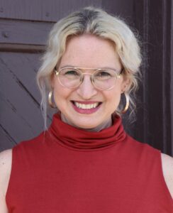 A white woman with blonde hair tied back in a red sleeveless turtleneck wearing glasses and gold hoop earrings in front of a dark gray wall.