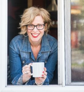 A white woman with short blonde hair leaning out a window holding a white coffee cup. She is wearing glasses and a blue long-sleeved shirt.
